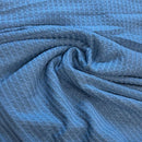 A close-up of textured blue fabric with a waffle pattern, arranged in soft folds and swirls.