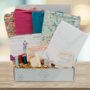 Crafting kit with fabric, thread, and stationery items in a box on a wooden surface.